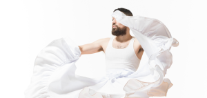 Image: Detail of a promotional photo from "World After This One," an experimental dance performance by Benji Hart. Hart is captured in movement, wearing a white tank top and skirt. The skirt is flung upward, covering half of their face. Photo by William Frederking.